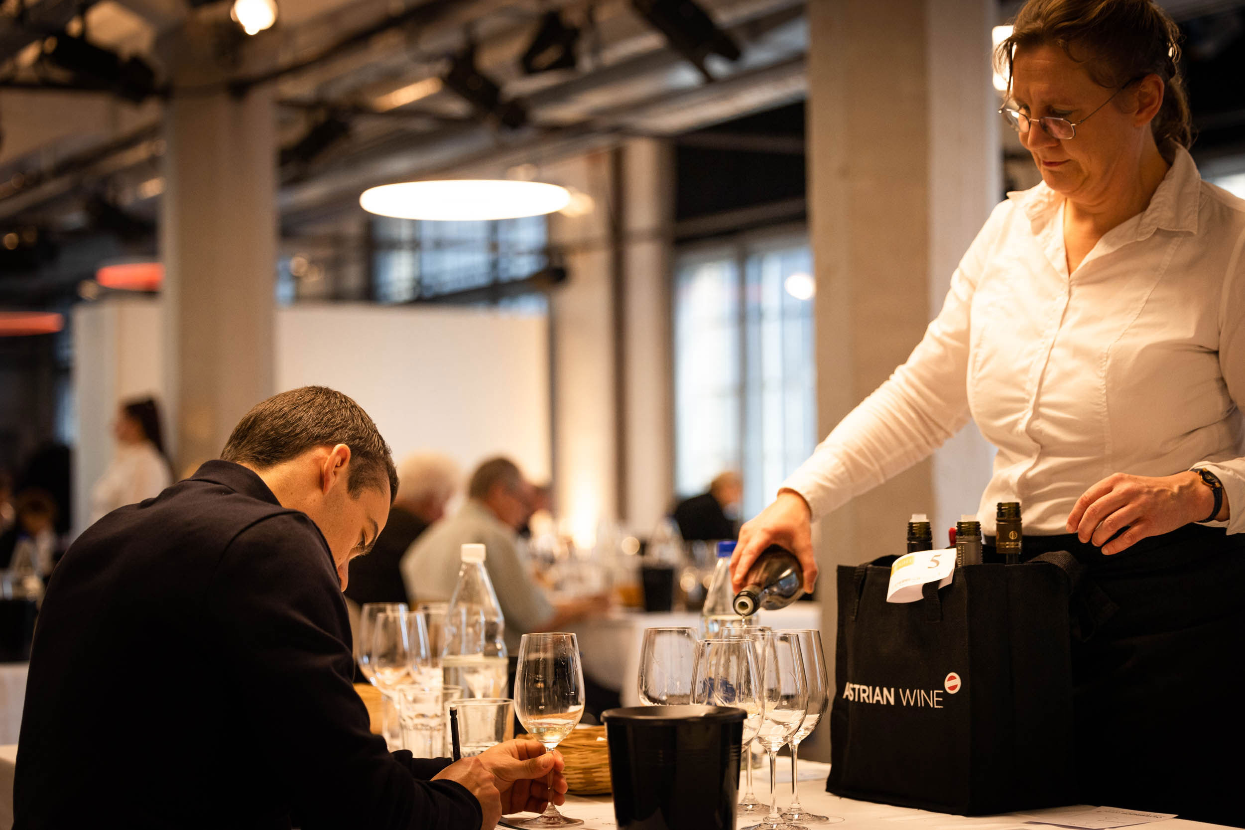 servicemitarbeiterin schenkt einem teilnehmer wein ein. er schreibt seine notizen nieder. vor der servicemitarbeiterin hat es eine tasche mit 6 verschiedenen Weinen, die einen Flight repräsentieren.