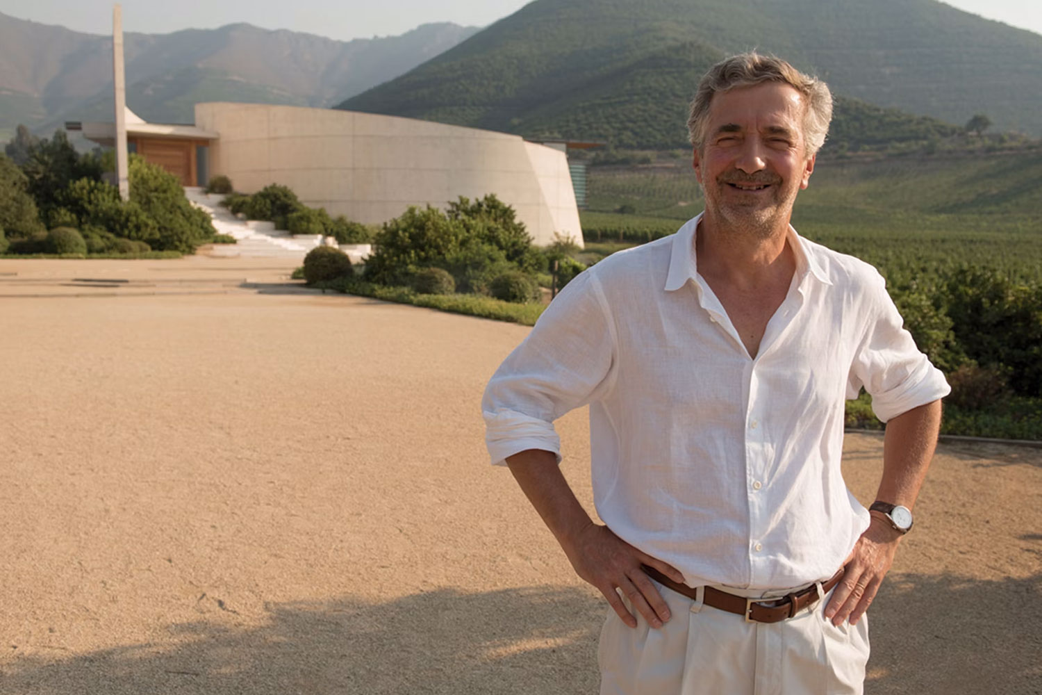 Eduardo Chadwick steht lächelnd im Freien vor einer modernen architektonischen Kellerei, umgeben von grünen Hügeln und Weinbergen.