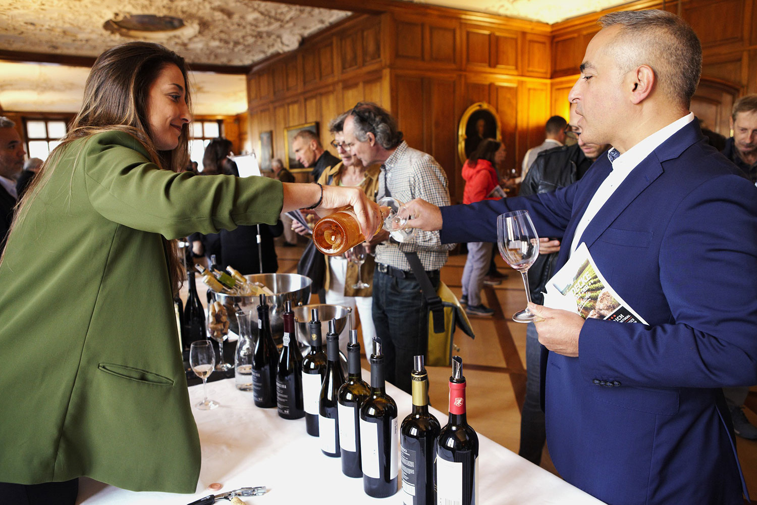 Eine Frau in einem grünen Blazer schenkt einem Mann in einem blauen Anzug ein Glas portugiesischen Wein ein. Sie stehen an einem Tisch mit mehreren Weinflaschen aus Portugal in einem eleganten Raum voller Menschen