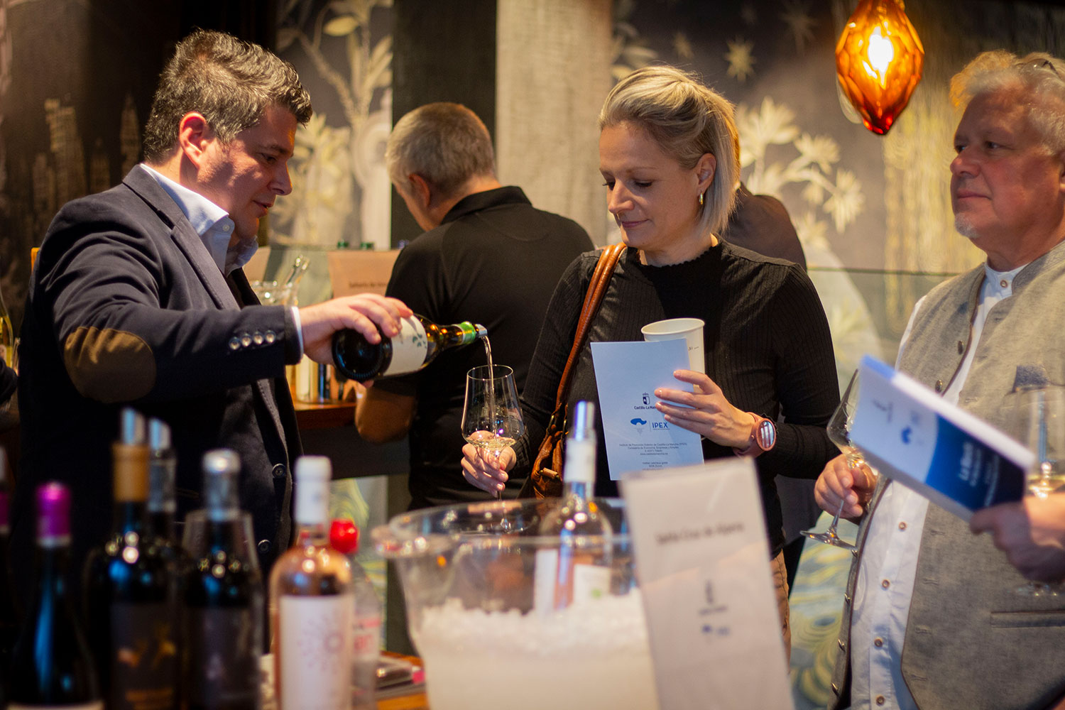 Eine La Mancha Weinverkostungsszene, bei der ein Mann in einem dunklen Sakko Wein in ein Glas einschenkt, während eine Frau mit einem Verkostungsheft in der Hand zuschaut. Im Hintergrund sind weitere Teilnehmer der Verkostung zu sehen.