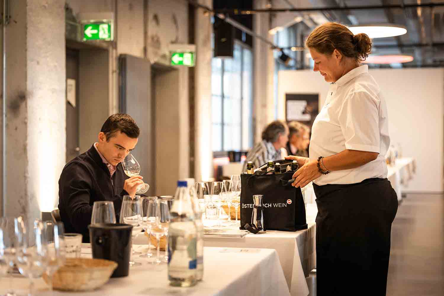 Weindegustation mit einer Person, die sorgfältig ein Glas Wein prüft. Im Hintergrund steht eine Servicemitarbeiterin mit einer Tasche, die mit 'Österreich Wein' beschriftet ist, was auf eine Verkostung österreichischer Weine hinweist. Auf dem Tisch sind mehrere Weingläser und Flaschen angeordnet, was zur Atmosphäre der Degustation beiträgt.