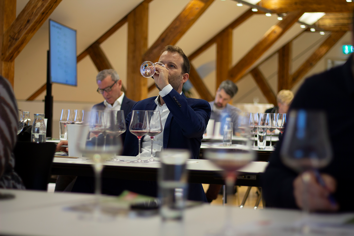 Teilnehmer einer Wein-Masterclass, der konzentriert ein Glas Rotwein probiert. Im Hintergrund sind weitere Personen in die Verkostung vertieft. Die Veranstaltung findet in einem stilvollen Raum mit Holzbalken statt, passend zur Atmosphäre einer professionellen Weinverkostung, mit Weinen aus Südtirol und der Ätna-Region. Mehrere Weingläser stehen auf den Tischen, die eine Vielfalt an Weinen für die Teilnehmer präsentieren.