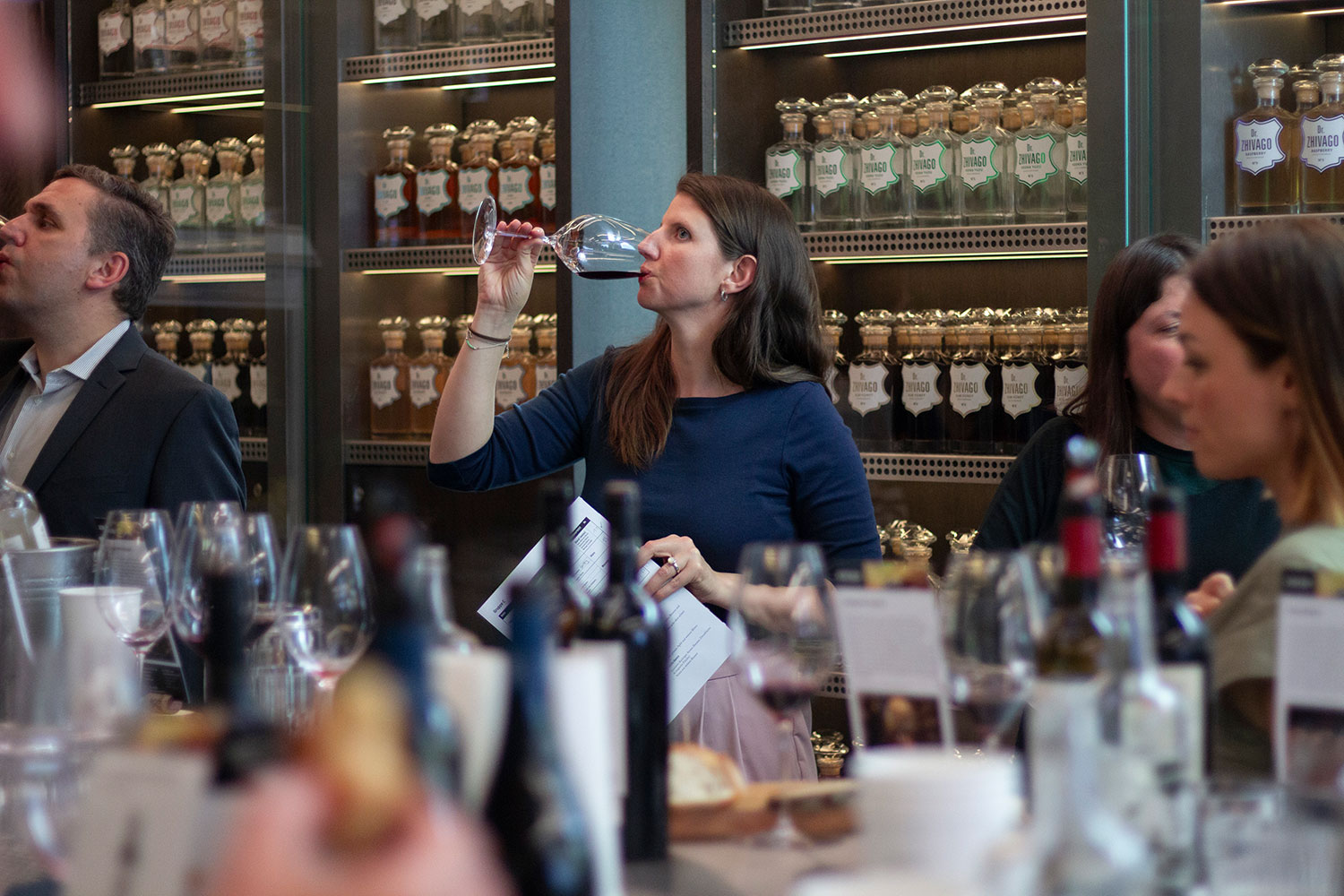 Teilnehmerin des Rioja Mates Events verkostet ein Glas Rioja-Wein. Im Hintergrund sind weitere Gäste und Flaschen sowie Regale mit dekorativen Flaschen zu sehen. Der Fokus liegt auf der Weinverkostung und dem Austausch unter den Rioja Mates.